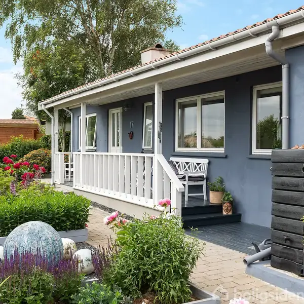 Hus med blå facade og hvid veranda, omgivet af en frodig have med blomster og en stor sfærisk dekoration. En sort komposter står ved siden af verandaen.