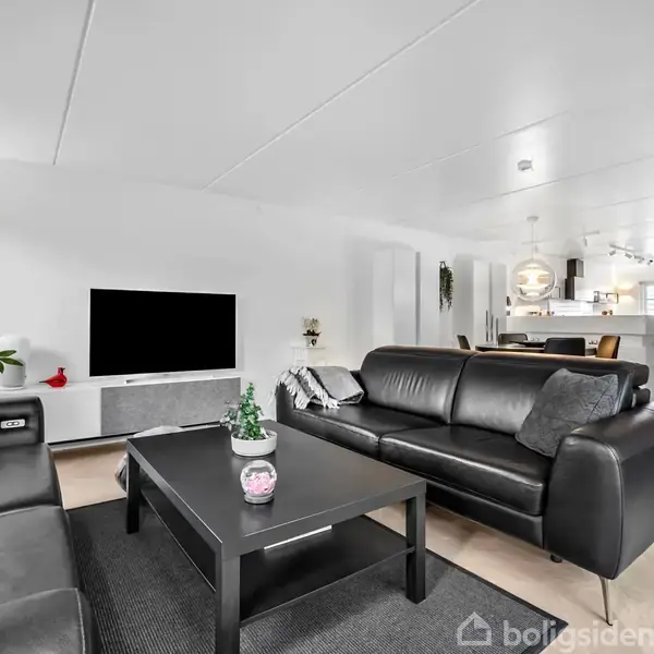 En moderne stue med sorte lædersofaer og et sofabord, der står foran et stort TV. Rummet er lyst og rummeligt, med en åben forbindelse til et køkkenområde.