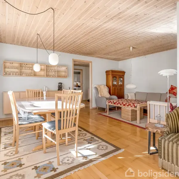 Spisebord med træstole står på tæppe i en stue. Sofa og lænestol findes i baggrunden. Træloft og trægulv skaber en varm atmosfære.