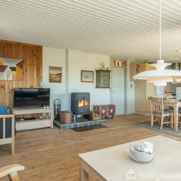 Stue med brændeovn, der opvarmer rummet. Der er en sofa og et tv ved en trævæg. Et spisebord og moderne lampe står ved siden af det åbne køkkenområde.