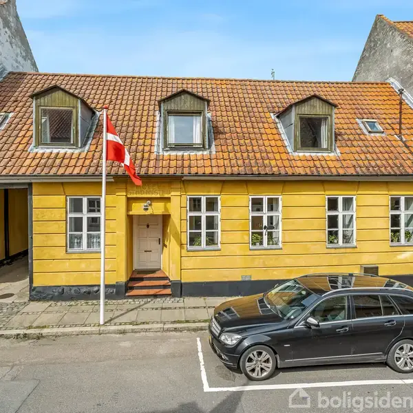 Et gult, etplans hus med rødt tegltag har tre kviste og fire vinduer foran. En bil er parkeret foran huset på gaden. En dansk flagstang står ved siden af hoveddøren.