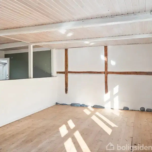 Et lyst rum med trægulv har en halv væg, der adskiller det fra et køkkenområde. Væggene er hvide med synlige bjælker. Store vinduer kaster lysskygger på gulvet.