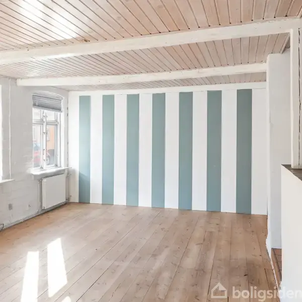 Værelse med lyse trægulve, der har tre vinduer til venstre og en væg med lodrette blå og hvide striber. Loftet er træpaneler, og der er radiatorer under vinduerne.