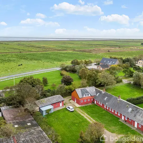 Røde bygninger med hvide vinduer og tage omgiver en græsplæne, hvor en hvid bil står parkeret. Bygningerne ligger på landet, omgivet af marker og spredte træer.