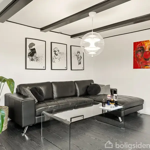 Sort lædersofa står i stue, foran glasbord med drikkevarer. Vækst til venstre, vægdekorationer, inklusive farverigt portræt til højre. Moderne loftslampe hænger over bordet.