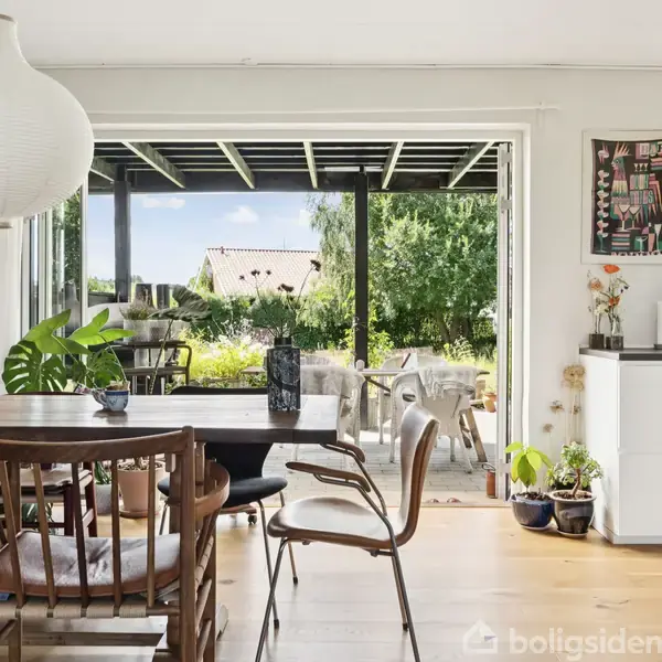 Spisebord omgivet af stole står i et lyst rum. En stor glasdør åbner ud mod en terrasse med udsigt til grønne træer og blå himmel. Plantepotter dekorerer hjørnerne.
