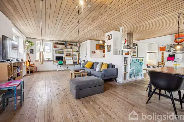 Stue med grå sofa og sofabord, omgivet af trævægge og trægulve. Et gyngesæde hænger fra loftet; skrivebord og bogreoler i baggrunden. Køkken med spisebord til højre.