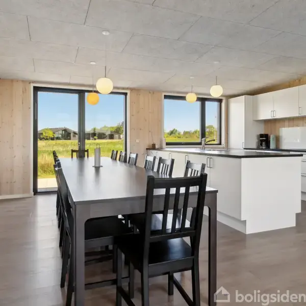 Langt spisebord med sorte stole står i en lys, moderne spisestue med store vinduer, der åbner ud mod en grøn udsigt. Køkkenet er placeret i det fjerne hjørne.