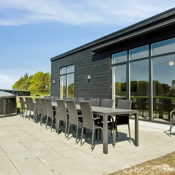 Et sort sommerhus har udendørs terrasse med lang række sorte stole og borde. I baggrunden ses et rundt boblebad. Terrassen er omgivet af grønne områder og træer.