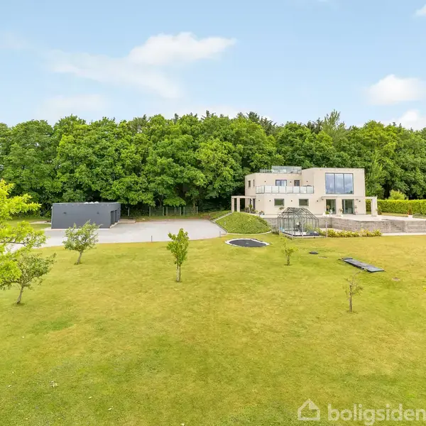 Et moderne hus står på en stor grøn græsplæne med spredte træer. En skov fungerer som baggrund, og en flisebelagt terrasse ses ved siden af huset.