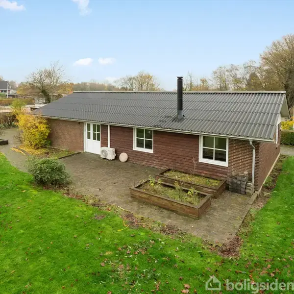 En étplans murstenshus med sort tag ligger på en stor, grøn græsplæne. Der er to hævede plantebede ved indgangspartiet. Omkring er der træer og en lille vej.