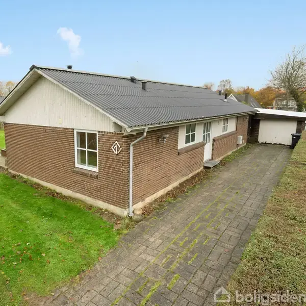 En brun murstensbungalow med gråt tag står ved en brolagt indkørsel. Huset er omkranset af velplejede grønne græsplæner og buske, mens træer kan ses i baggrunden.