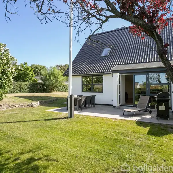 Et hus med sort tag står i en solrig have. På terrassen foran huset står havemøbler og liggestole, omgivet af grønne buske og træer under en klar blå himmel.