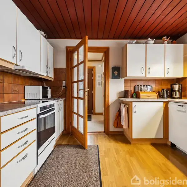 Et køkken med hvide skabe og trædetaljer, fyldt med apparater som ovn, mikrobølgeovn og kaffemaskine. Køkkenet har brun flisevæg, træloft og en åben dør til en gang.