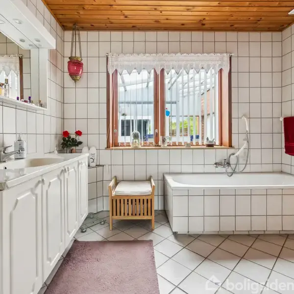 Et badeværelse med hvidt flisegulv og vægge har en hvid håndvask, et badekar og et vindue med udsigt til en veranda. En rød håndklæde hænger ved siden af badekarret.