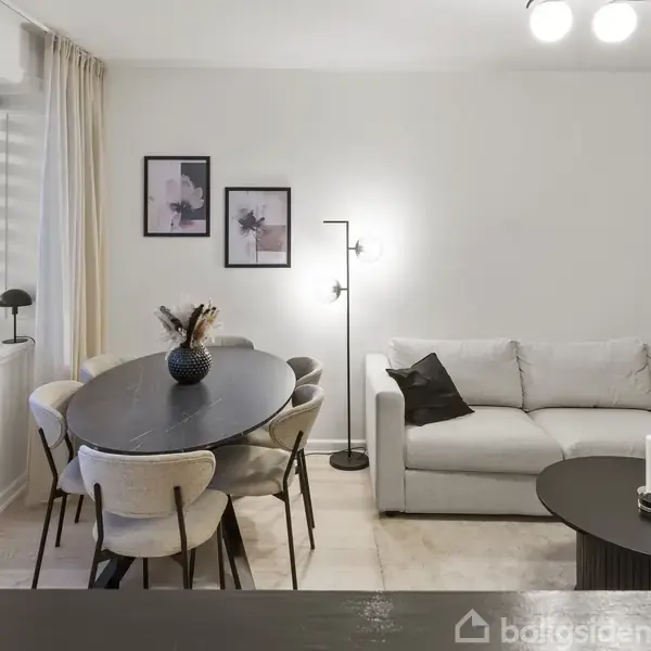 En stue med en grå sofa og moderne lamper; et rundt spisebord med seks stole står til venstre. Væggene har minimalistisk kunst, og vinduet har lyse gardiner.