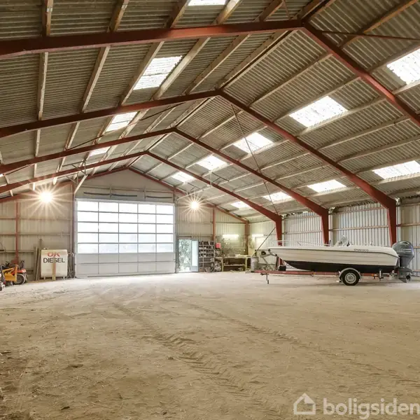 En stor maskinhal med en båd placeret på en trailer. En person arbejder nær båden. Rummet er rummeligt med en jordgulv, stålsøjler og flere tagvinduer.