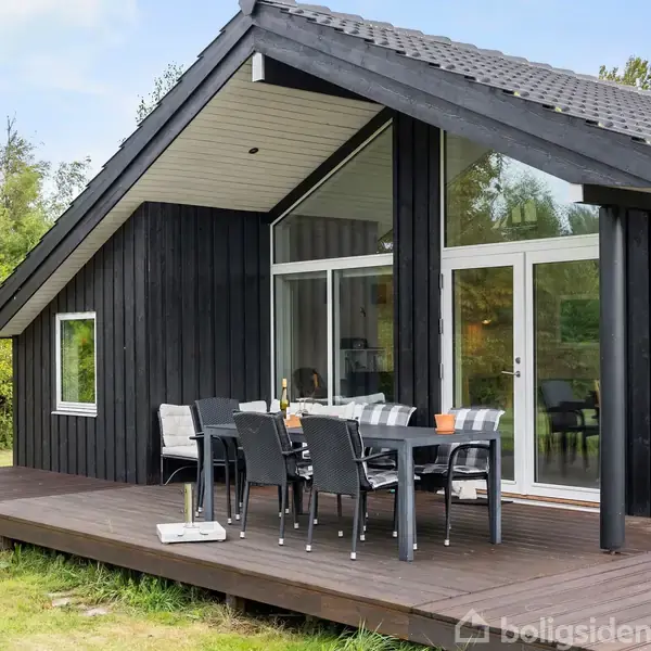 Et sort træsommerhus står stille på en træterrasse. Havemøbler er arrangeret foran store vinduespartier. Omgivet af grønne buske og skov, skaber det en rolig atmosfære.