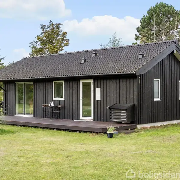 Et sort træhus står på en grøn græsplæne, omgivet af buske og træer. Huset har en træterrasse med et lille bord og to stole.