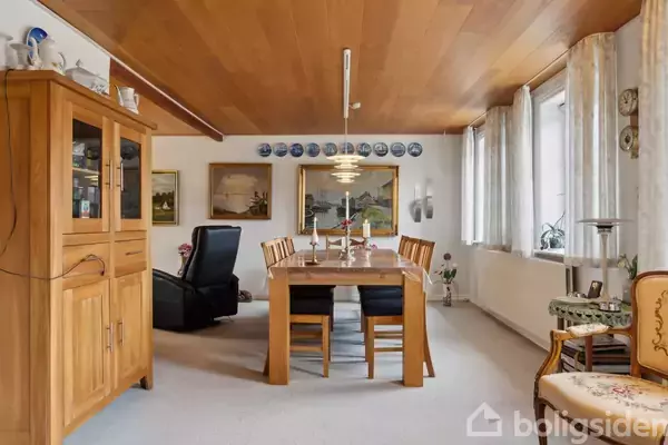 Et spisebord med seks stole står i en stue med træloft og lyse vægge. Et vitrineskab og dekorerede tallerkener hænger på væggen. Store vinduer til højre slipper lys ind.