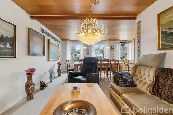 En stue indrettet med en sofagruppe og lænestol under en stor, dekorativ lysekrone. I baggrunden ses et spisebord omgivet af vinduer med udsigt over en have. Malerier pryder væggene.