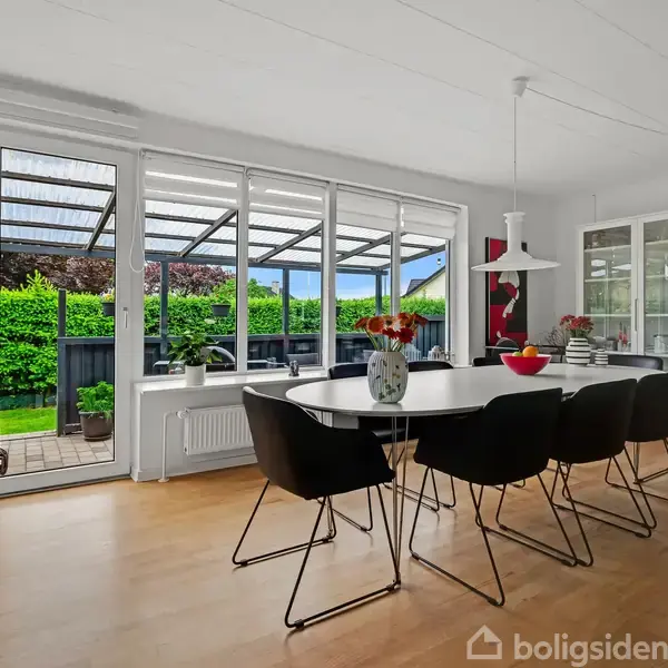 Spisebord med seks sorte stole står i en lys spisestue med glasdøre ud til en terrasse og have. Skabsreol med glaslåger til højre.