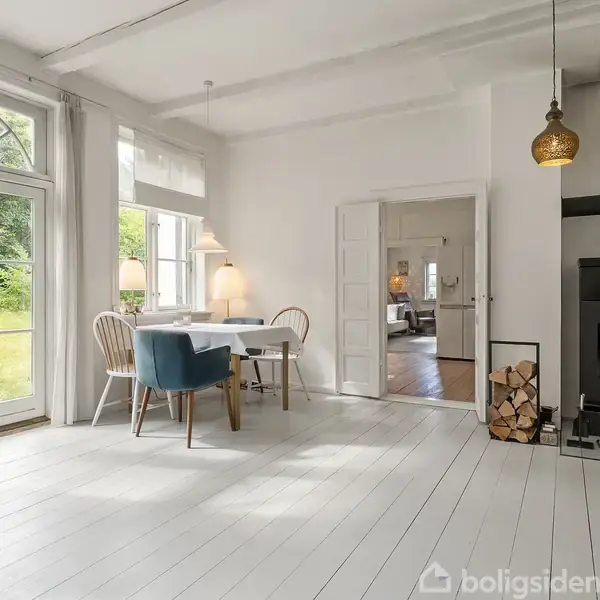 En moderne stue med en brændeovn tændt, ved siden af en stabel træ. Et spisebord med stole står ved et vindue ud til en grøn have.