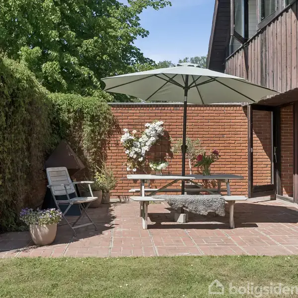 En terrasse med et bord og stole under en stor parasol. Omgivet af murstensvægge og planter, med udsigt til en have.
