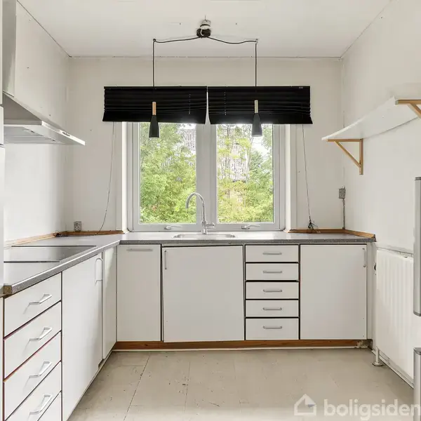 Et moderne køkken med hvide skabe, en indbygget ovn og et køleskab står i fokus. En vask er placeret under et stort vindue med udsigt til grønne træer.