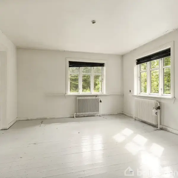 Et tomt rum med hvide vægge og to vinduer med sorte persienner. Rummet har et lyst trægulv og indeholder radiatorer under vinduerne. Udsigt til grønne omgivelser udenfor.