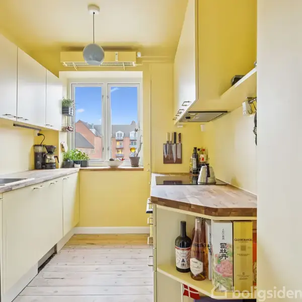 Et langt køkken med lysegule vægge og hvide skabe indeholder en bordplade med vinflasker samt en kaffemaskine. Et vindue i enden giver udsigt til nabobygninger.