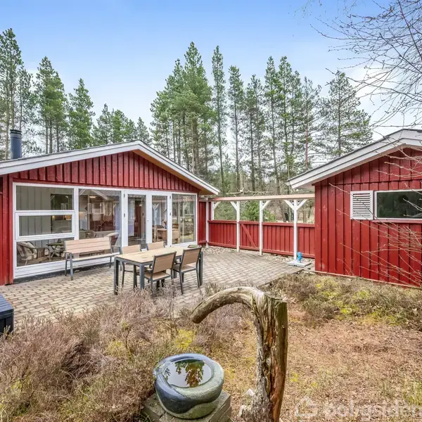 Rødt sommerhus med terrassebord og stole foran, omgivet af fyrretræer. En grill står ved siden af en bænk, og der er et fuglebad i forgrunden.