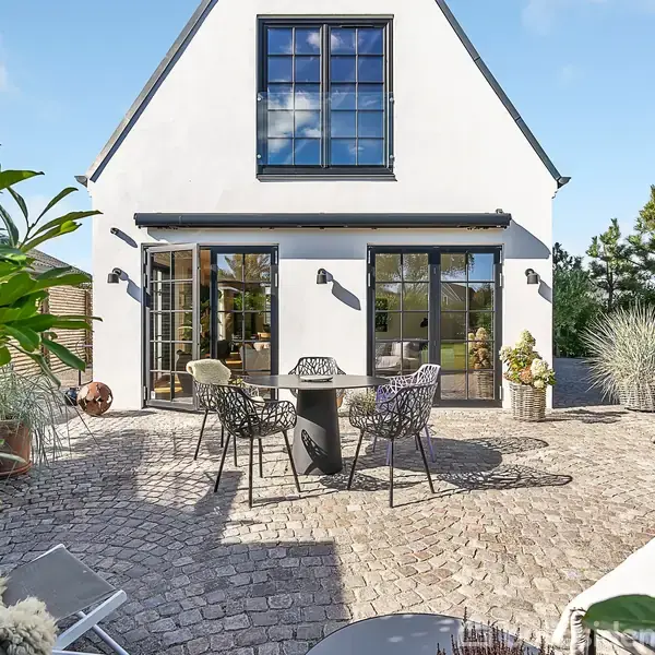 Hvidt hus med store glasdøre åbner ud til en brostensbelagt terrasse med et rundt bord og fire stole. Omkring terrassen ses frodig beplantning og pænt vedligeholdte buske.