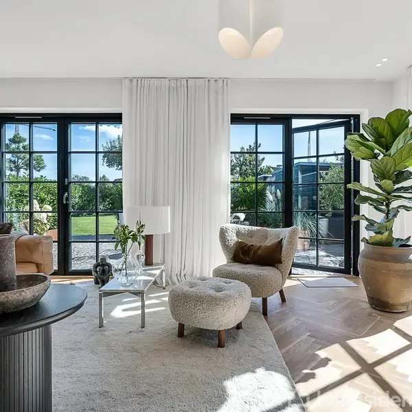 Stue med moderne indretning; en lysebrun lædersofa står ved siden af et rundt bord med dekorationer. Store vinduer lukker lys ind, og en grøn plante pynter hjørnet. Udsigt til have.