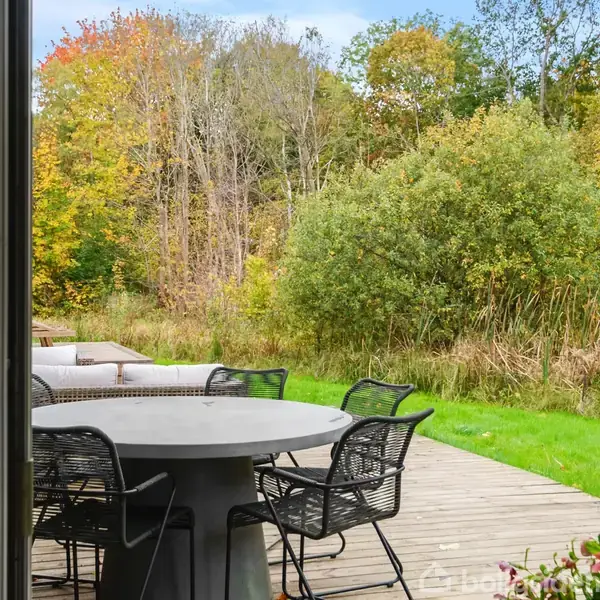 Rundt havebord med sorte stole står stille på en træterrasse. Bordet er omgivet af en græsplæne og tæt bevoksede træer i efterårsfarver.