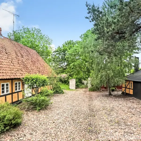 Bindingsværkshus står stille omringet af frodige træer og buske. Huset har gule vægge og rødt tegltag, mens gårdspladsen består af brosten. Et lysthus ses til højre.