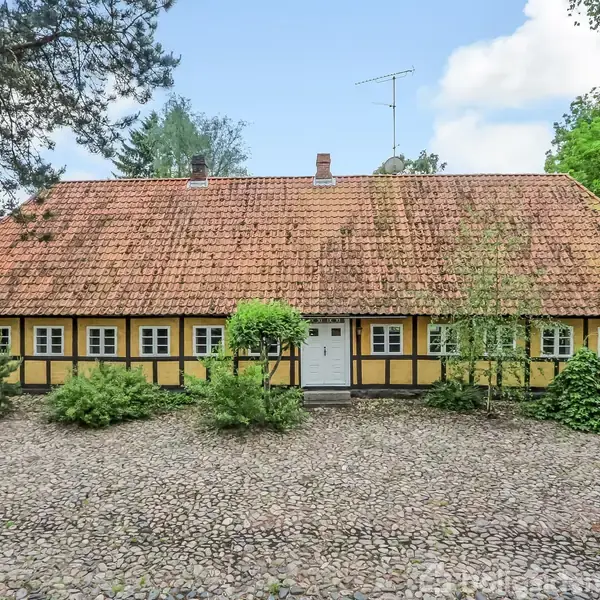 Hus med bindingsværk og rødt tegltag står stille omgivet af grønne træer og buske på en brostensbelagt gårdsplads under en blå himmel.