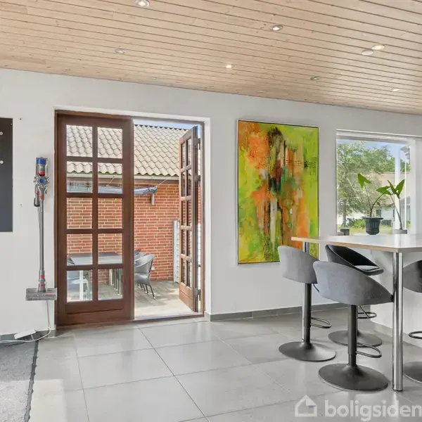 Et køkken med et hvidt køkkenbord og to grå stole. En fransk dør åbner ud til en terrasse. Et farverigt maleri hænger på væggen ved siden af et vindue.