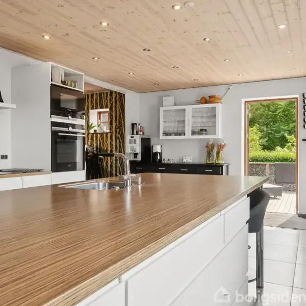 Køkken med en stor, træbeklædt ø, omgivet af moderne apparater og åbne hylder fyldt med krydderiglas. Gennemsigtig dør åbner ud til en terrasse med grøn beplantning.
