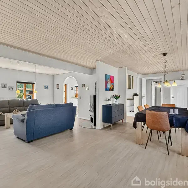 Stue med åben planløsning viser sofa og lænestole til venstre, spisebord med seks stole til højre under lamper. Lyse trægulve, hvidmalede vægge, og træloft udgør rummet.