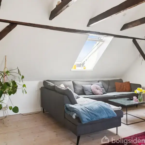 En grå sofa står under et skråt loft med synlige bjælker. Sofaen har puder og et tæppe. Ved siden af er et sofabord med blomster, og der er et maleri på væggen. En plante står ved siden af sofaen.