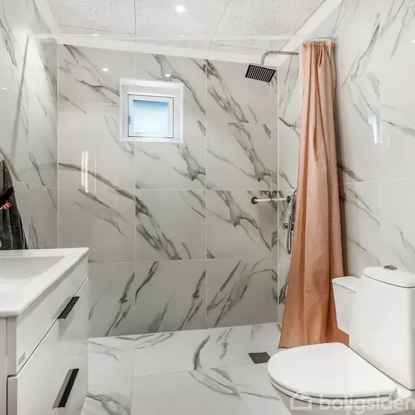 Badeværelse med marmorvægge og gulv. Toilet og håndvask er stationære, mens et brusehoved hænger i en bruseniche med et lyserødt forhæng. Et lille vindue er placeret over brusenichen.