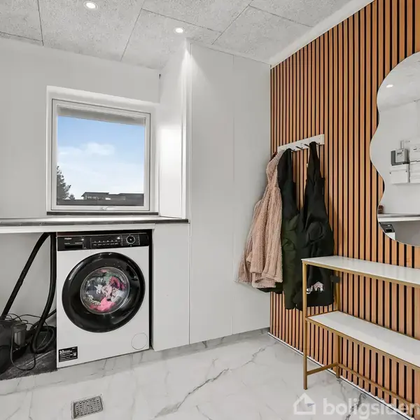 En moderne vaskerum med en vaskemaskine der vasker tøj, placeret under en bordplade ved et vindue. En stumtjener med jakker og en unik spejl hænger på trælamelvæggen.