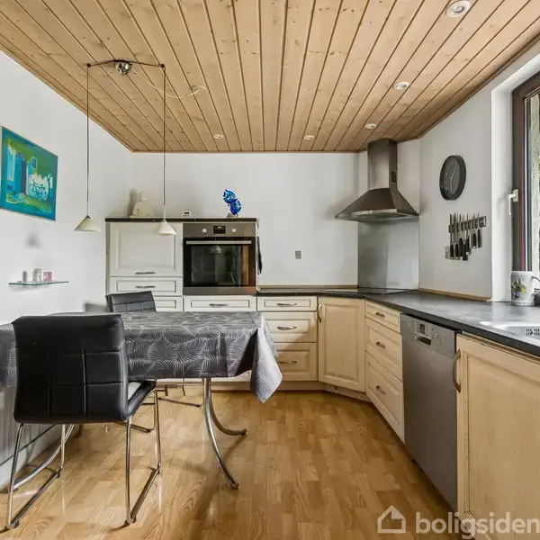 Køkkenbord er dækket med grå dug, omringet af stole. Lys træloft over. Indbygget ovn, opvaskemaskine og vask i enhed. Vindue åbner mod grøn udsigt. Ingen tekst til stede.