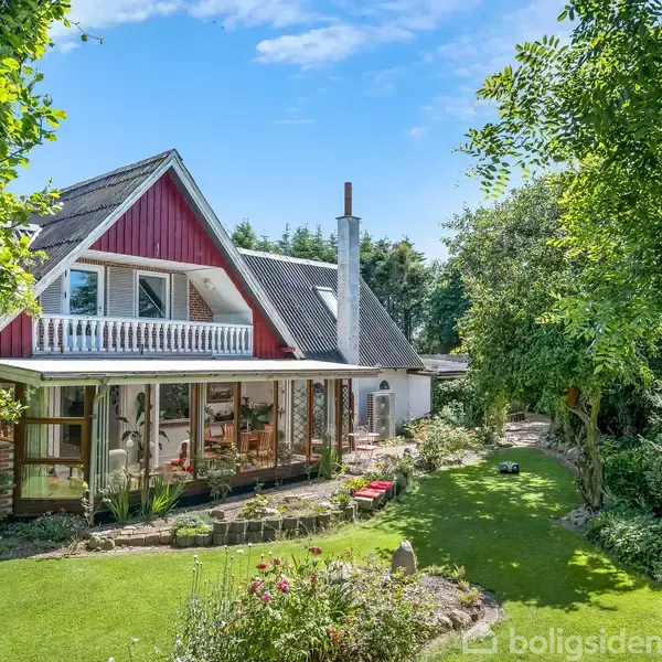 Huset, en rummelig to-etagers bygning med rødt og hvidt træværk, står i en frodig have fuld af træer og buske. Sollys oplyser den velplejede græsplæne.