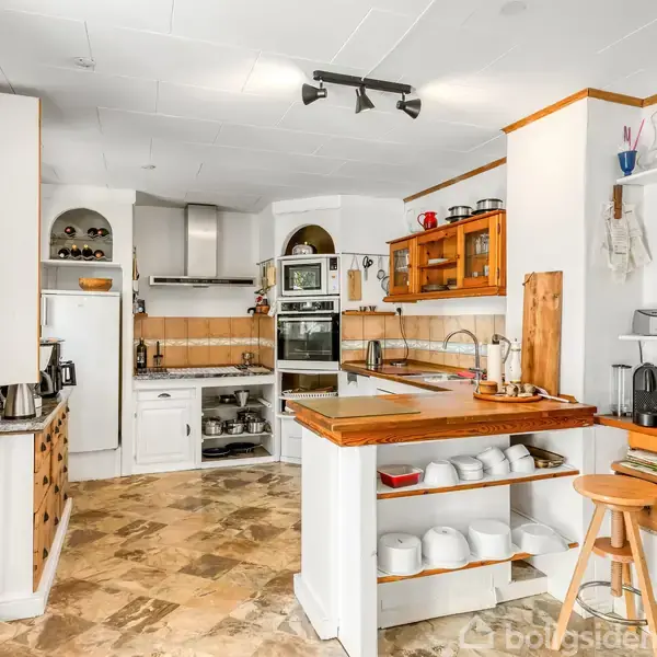 Et lyst køkken med marmorflisegulv, træhylder og en central ø fyldt med porcelæn. Hvidevarer indbygget i væggen, og der er forskelligt køkkentilbehør på hylder og bordplade.