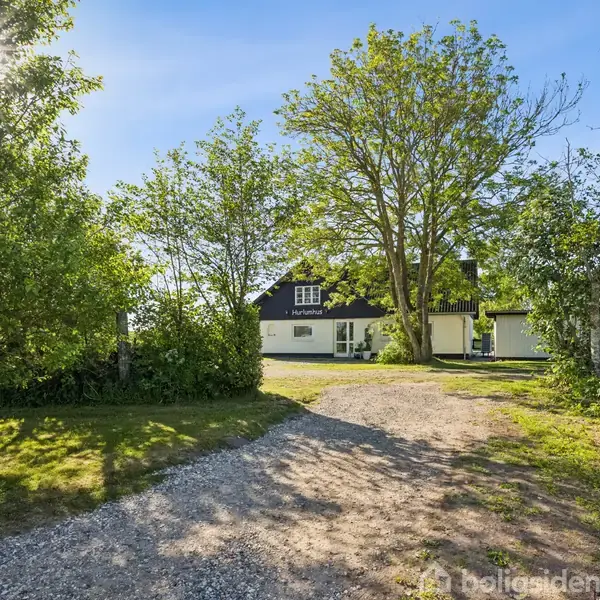 Hus beliggende bag en grusvej omgivet af træer og grøn vegetation. Sollys skinner gennem trætoppene, mens himlen er klar og blå.