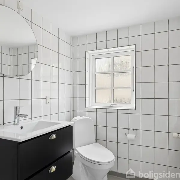 Spejl og vask står ved siden af et toilet i et moderne badeværelse med hvide flisevægge. Der er et vindue ved siden af toilettet og en radiator til højre.