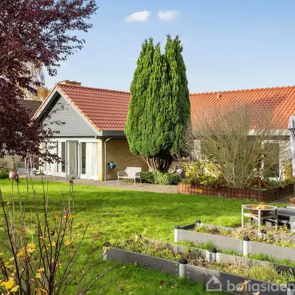 En enkeltfamiliehus med rødt tegltag ligger i en velholdt have. Derudover er der højbede og havemøbler med parasol placeret på græsplænen.