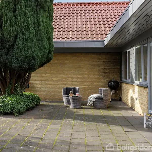 En terrasse med fliser og havemøbler ligger ved siden af et murstenshus. En stor busk står til venstre. Grillen står op ad væggen.